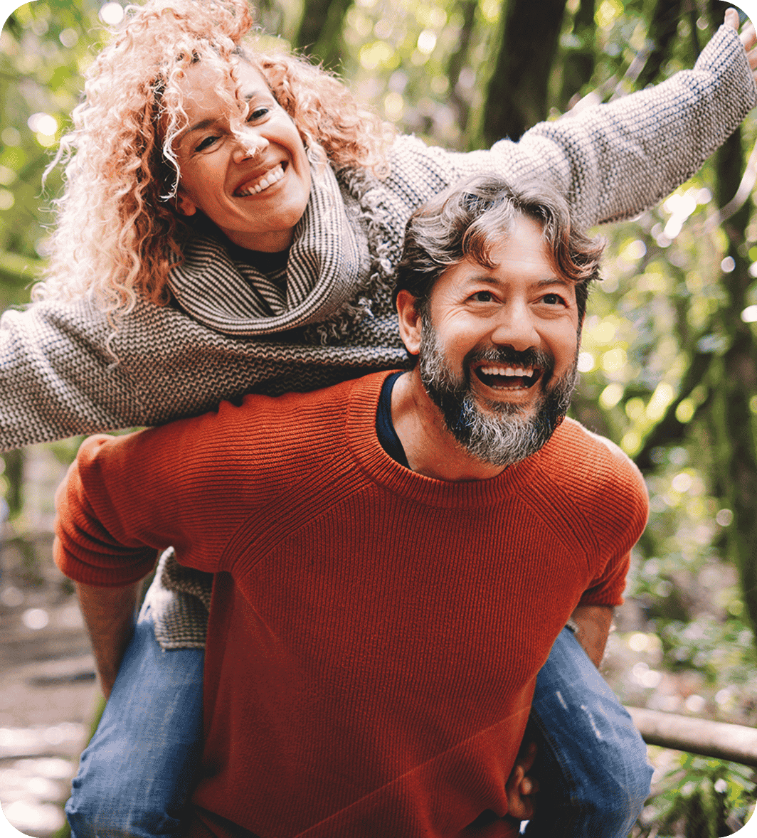 A man with a beard wearing an orange sweater gives a piggyback ride to a smiling woman with curly hair in a forest setting.