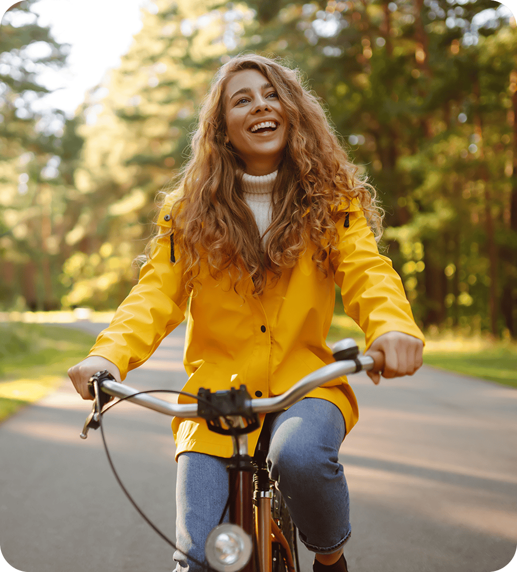 A woman with long curly hair smiles as she rides a bicycle on a paved path through a forest, wearing a bright yellow jacket and jeans.