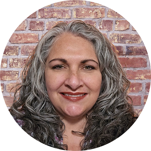 A person with curly gray hair smiles in front of a brick wall background.